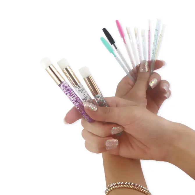 Two hands display makeup tools: one holds three glittery silicone brushes, the other six Apothie Disposable Lip Applicator Wands for precise makeup application. Both hands have glittery nail polish, and one wrist wears a beaded bracelet.