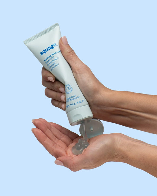 A person squeezes clear, firm-hold styling gel from a white tube labeled Aquage Molding Megagel 4 oz into their palm against a light blue background.