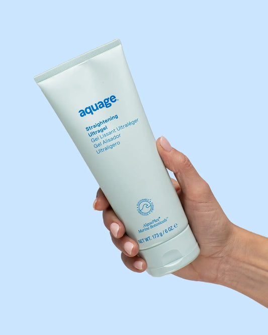 A hand holds an Aquage Straightening Ultragel 6 oz tube against a light blue background. The tube displays product details and provides humidity resistance for smooth, manageable hair.