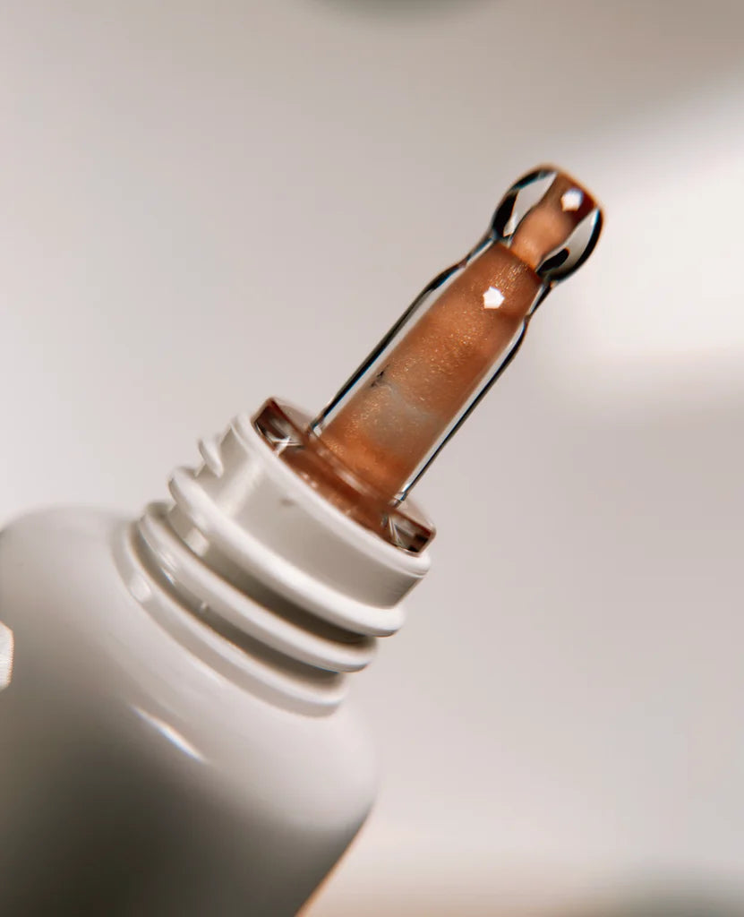 A close-up of Bodyography Radiant Glow Primer + Niacinamide’s dropper, filled with shimmery bronze primer and attached to a white bottle, against a soft neutral background—ideal for achieving a luminous, hydrated glow.