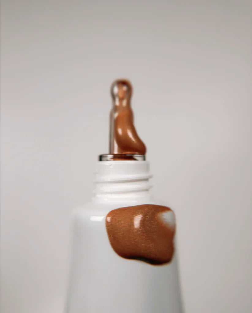 A close-up of the Bodyography Radiant Glow Primer + Niacinamide shows its white tube with metallic applicator tip dispensing shimmery bronze primer, with some of its niacinamide-infused formula smeared beside it on a plain background.
