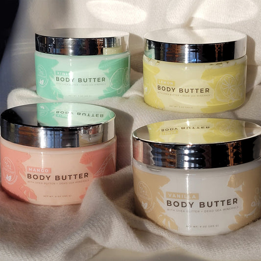 Four jars of Natural Elephant Dead Sea Shea Body Butter 9 oz in kiwi, lemon, mango, and vanilla are displayed on white fabric. Each has a silver lid and pastel label, enriched with Dead Sea minerals and plant oils for hydration.