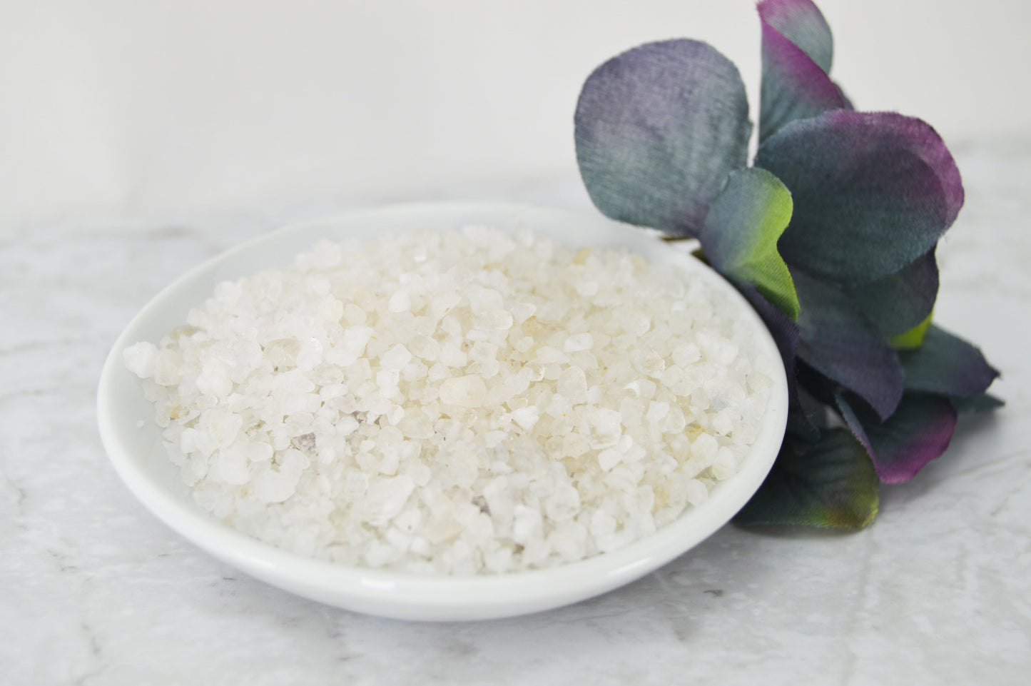 A white bowl filled with Natural Elephant Lavender Dead Sea Salt rests on a marble surface beside a purple and green artificial flower, perfect for enhancing your relaxation routine.