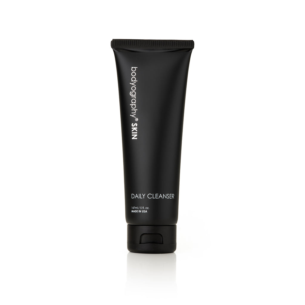 A black tube of Bodyography Skin Care Daily Cleanser 5 oz, featuring glycolic acid and free from sulfates and parabens, stands upright on a white background. The label lists product details and its 5 oz (140 ml) volume.