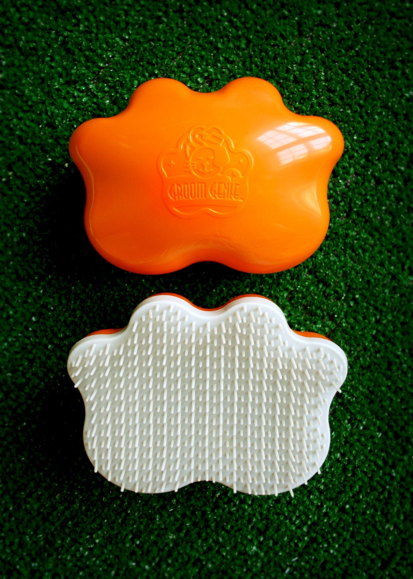 The Groom Genie Original Pet Detangling Brush, featuring an orange paw-shaped top and distinctive bristles, rests on its white underside atop a textured green artificial grass surface.