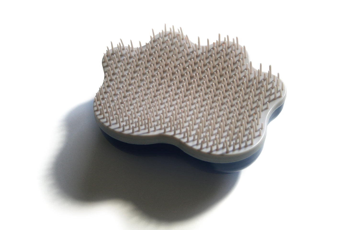 A close-up photo shows the Groom Genie Original Pet Detangling Brush, cloud-shaped in white with many short, flexible bristles and a unique design, casting a shadow on a white surface—ideal for innovative pet grooming.