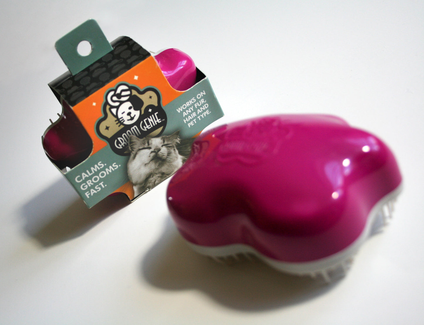 A pink and white Groom Genie Original Pet Detangling Brush featuring a unique bristle design sits next to its box, which displays a cat illustration and the phrases “Calms. Grooms. Fast.” and “Works on any fur, any pet type.”.