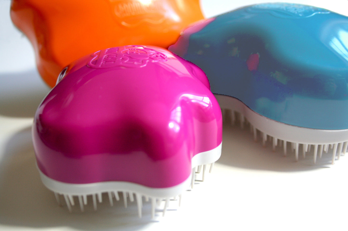 Three vibrant plastic hairbrushes—pink, blue, and orange—are shown close up. With shiny curves and white bristles, their unique design is inspired by the Groom Genie Original Pet Detangling Brush. The brushes overlap on a white surface.