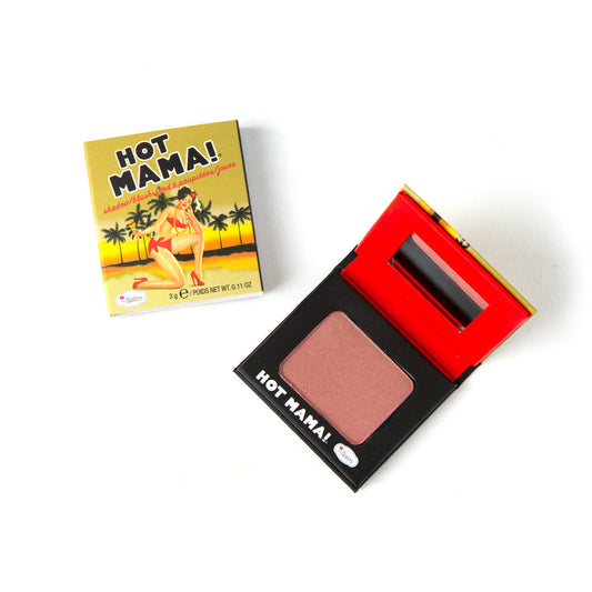 An open theBalm Hot Mama Travel Size Blush compact shows pink, shimmery blush and a small mirror. Nearby, the closed box with vintage pin-up art and palm trees sits on a white background.