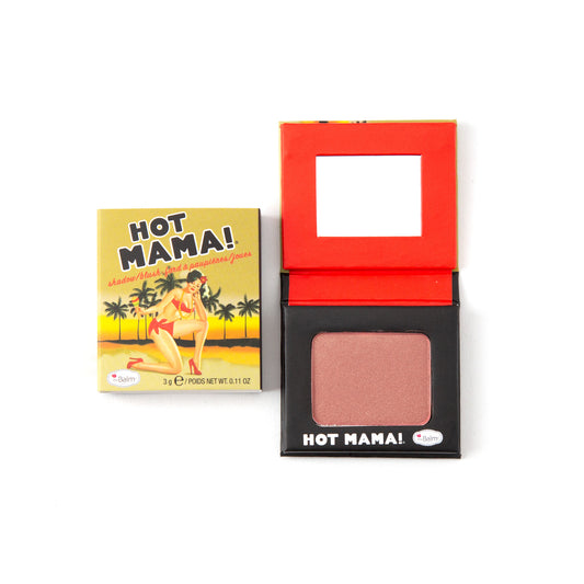 An open compact of theBalm Hot Mama Travel Size Blush sits beside its closed packaging, which shows a vintage-style woman in a bikini with palm trees and a sunset, emphasizing the blush’s subtle shimmer.
