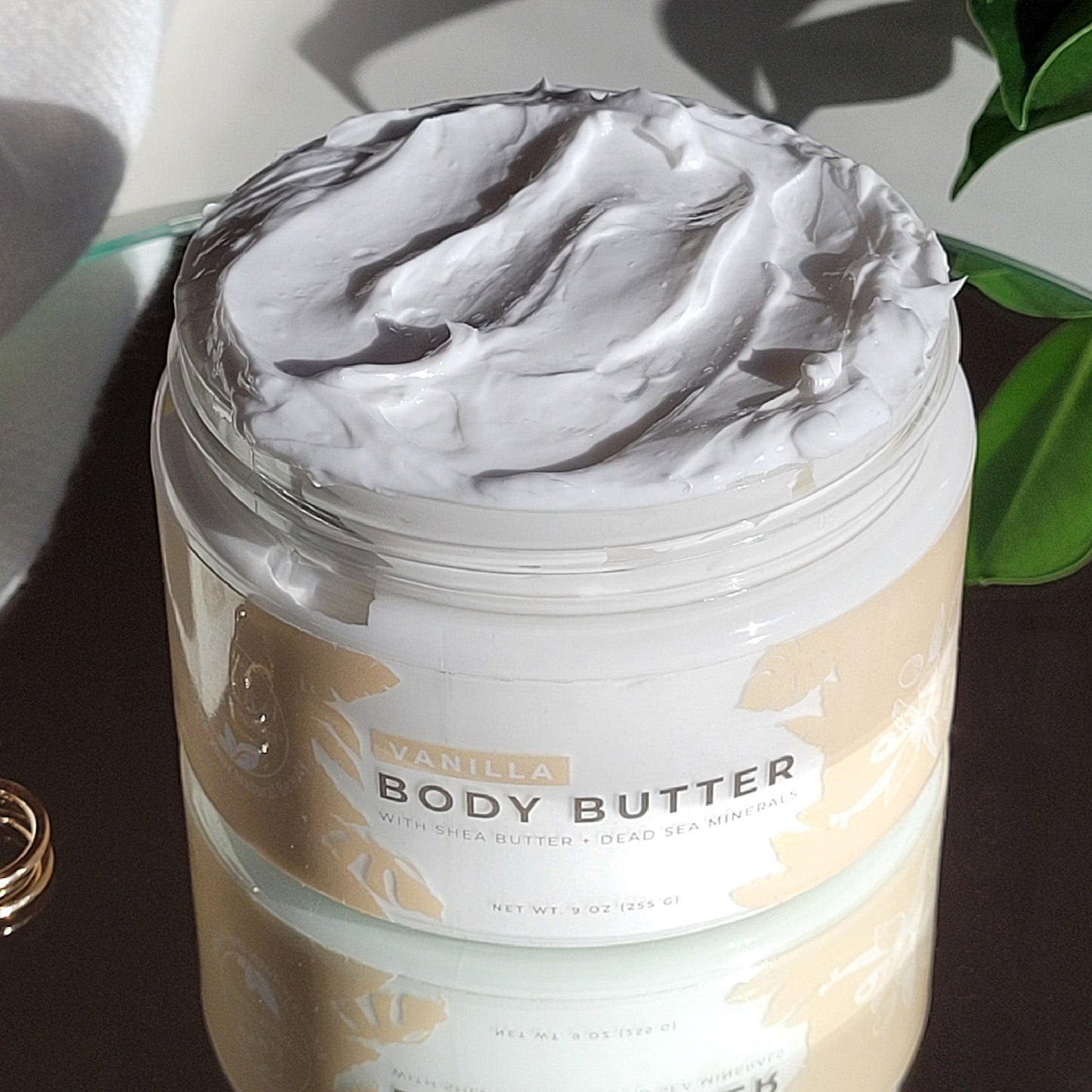 An open jar of Natural Elephant Dead Sea Shea Body Butter 9 oz reveals its creamy, whipped texture. The label emphasizes natural plant oils, shea butter, and Dead Sea minerals. The jar rests on a reflective surface with greenery nearby.