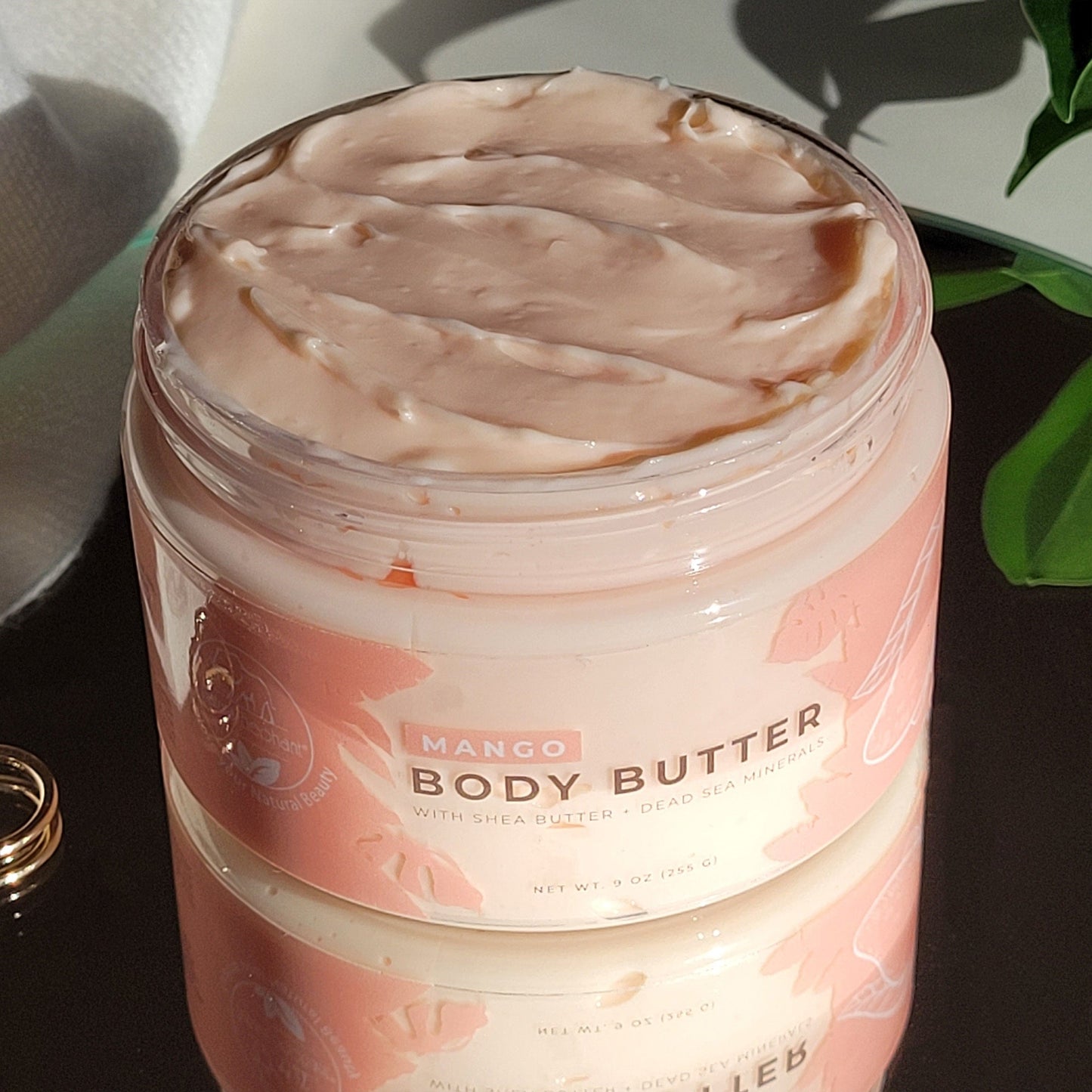 An open jar of Natural Elephant Dead Sea Shea Body Butter 9 oz displays its creamy texture, with the label visible. The jar is reflected on a glossy surface, accompanied by a plant and a ring nearby.