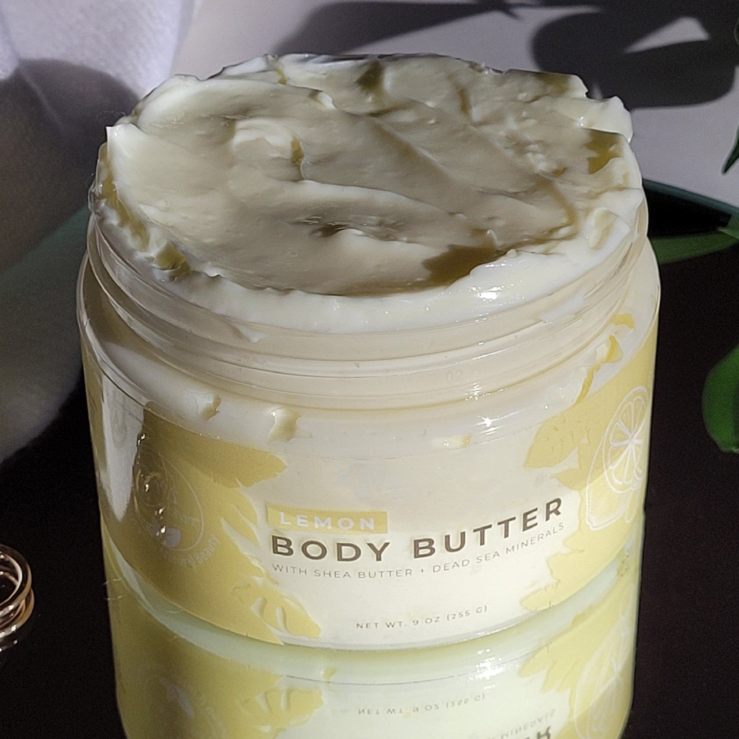 An open jar of Natural Elephant Dead Sea Shea Body Butter 9 oz rests on a surface, revealing its creamy texture. The label includes yellow accents and lemon illustrations.