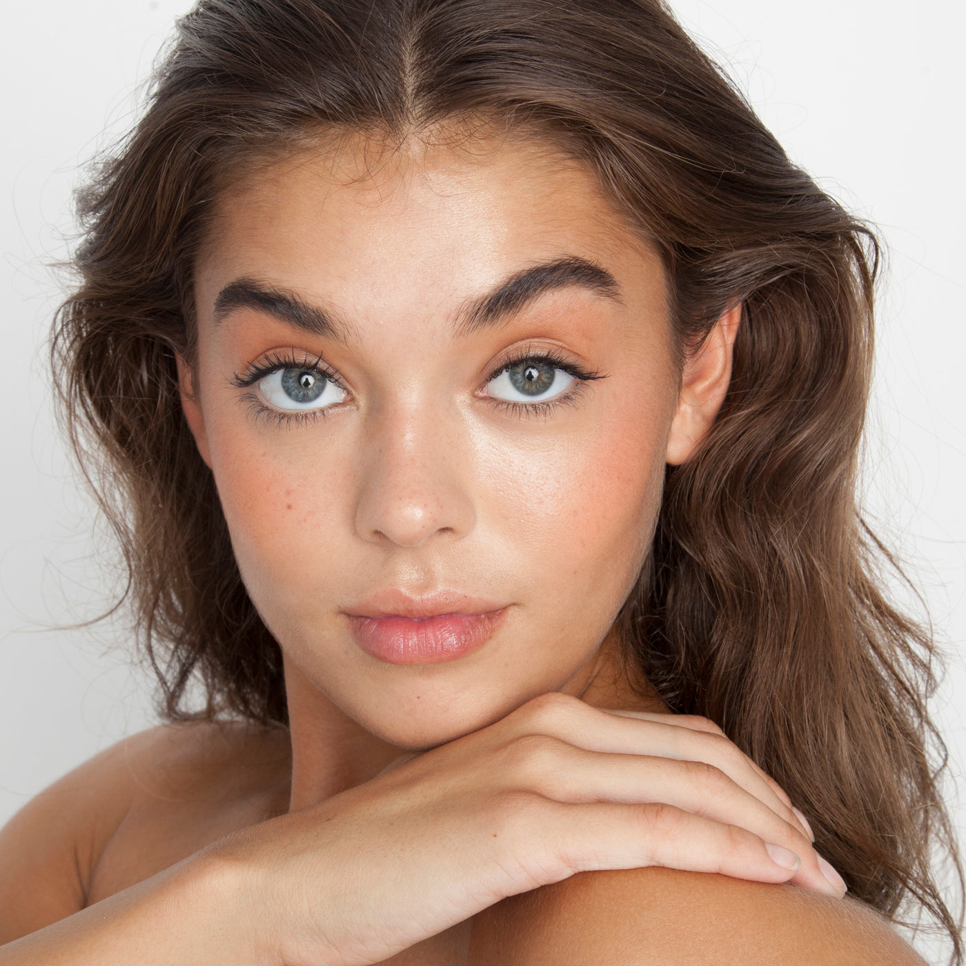 A young woman with wavy brown hair and blue eyes looks at the camera, her hand on her shoulder. She wears natural makeup with defined brows, long lashes, a neutral lip color, and theBalm Hot Mama Travel Size Blush against a white background.