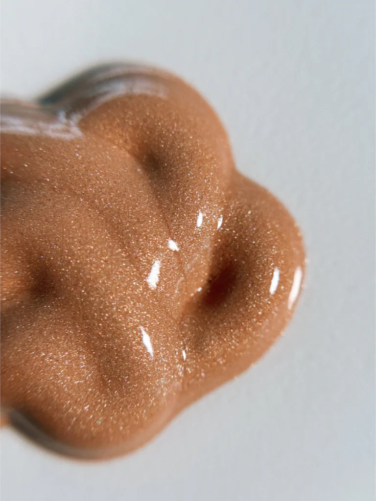 A close-up of Bodyography Radiant Glow Primer + Niacinamide, a hydrating, bronze-gold liquid with a shimmery metallic sheen and radiant glow, spread in an irregular shape on a smooth, light-colored surface.