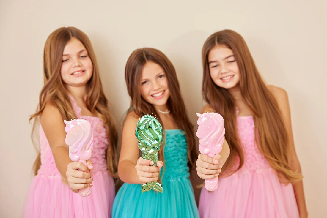 Three girls in colorful dresses holding unicorn detangling hair brushes with joyful expressions