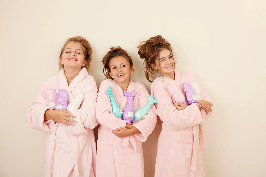 Three girls in pink robes happily holding unicorn conditioner for kids in colorful bottles