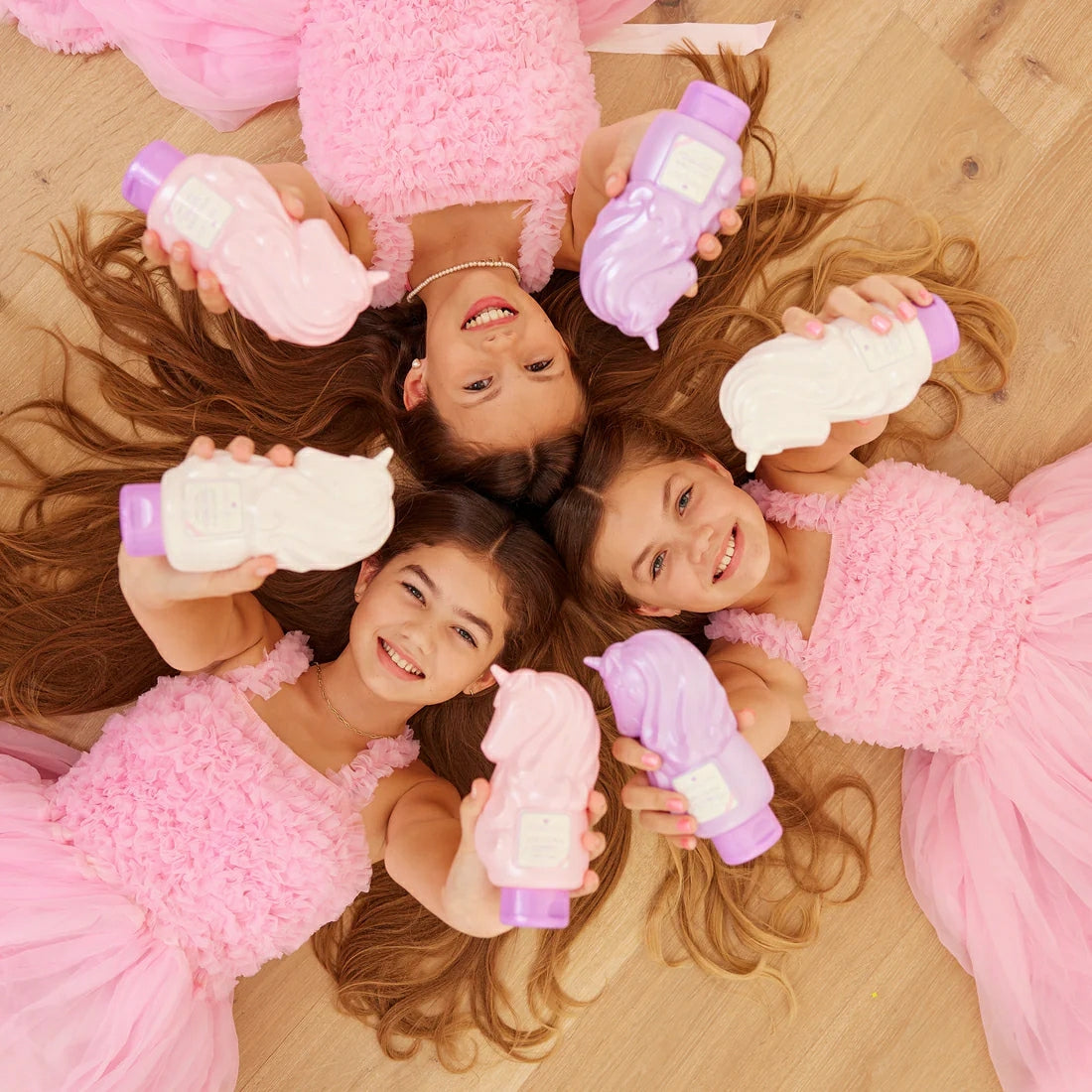 Three girls in pink dresses holding Glimmer Wish unicorn shampoo for kids, smiling and having fun