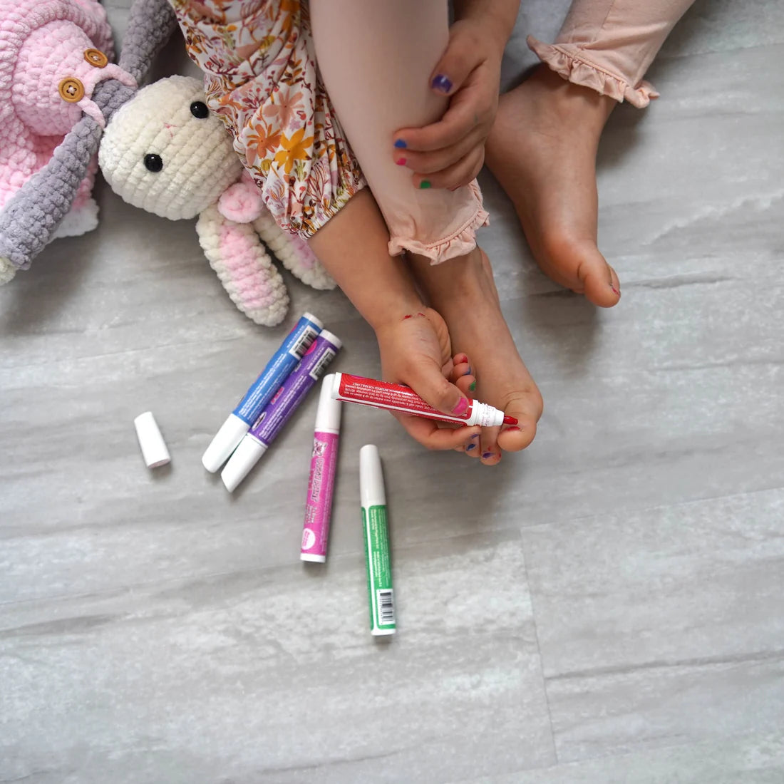 Child using Piggy Paint pen polishes on toes while playing with plush toys on the floor