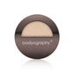A round metallic compact of Bodyography Highlighter Pressed Powder Sunsculpt Duo - Bronzer/Highlighter features a transparent window showing a shimmery beige shade, with the brand name bodyography printed in white on the lid.