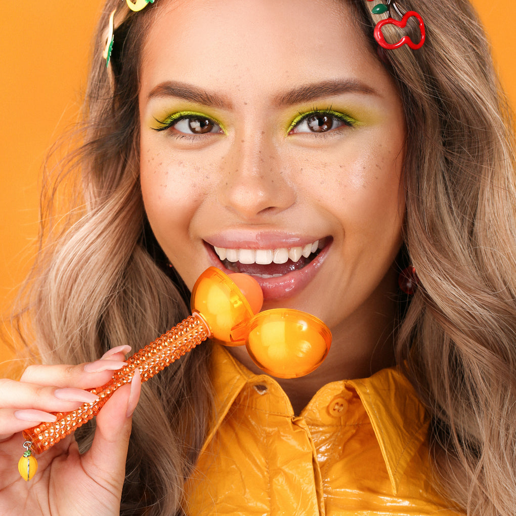 Model holding Glossy Pops TGI Fruity lip balm in vibrant orange with playful accessories
